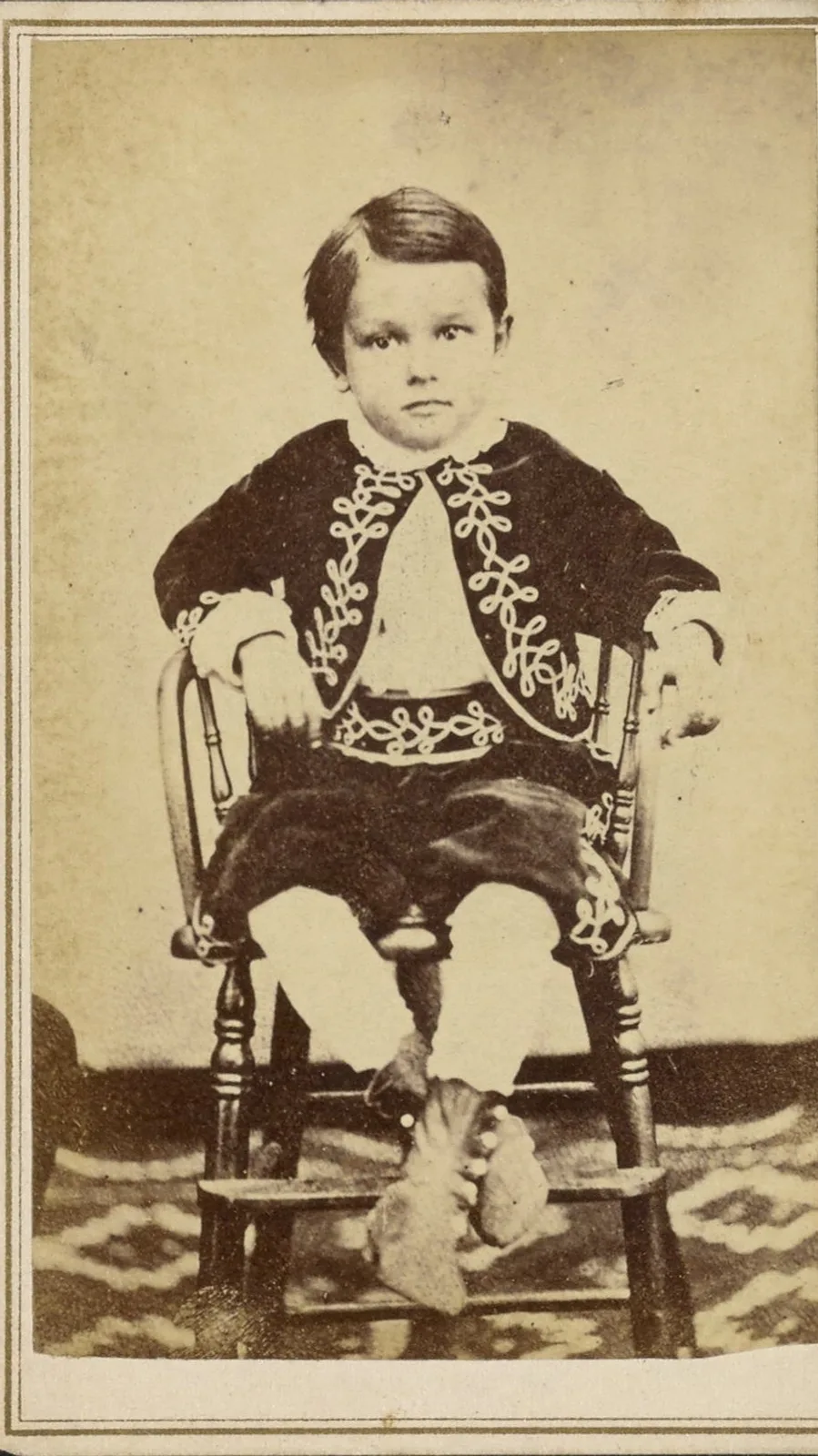 Original: A small boy in an embroidered jacket seated on a wooden chair, cabinet card